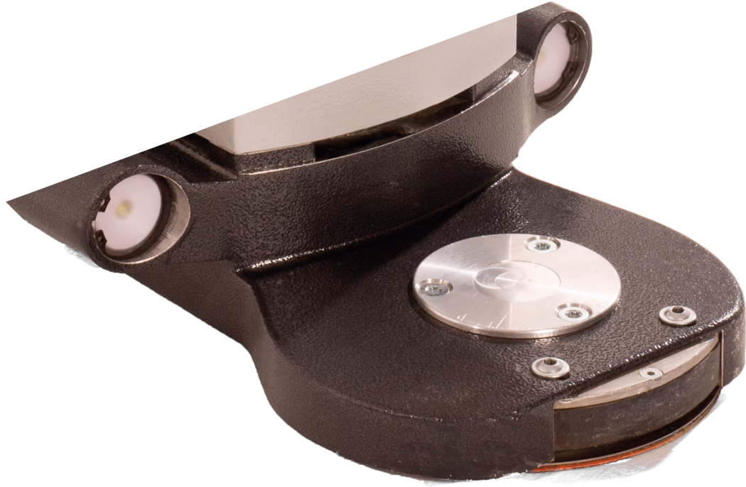 PALLMANN_dummy_Gecko Star attachment length short_msl_2018-07 Close-up of a small industrial electromechanical component photographed on a white background. The object features a textured dark metallic housing with curved contours, a polished flat metal contact or plate at the top and a round machined silver disc secured by three small screws on the upper face. Two circular recessed LED-like modules or indicator lenses with white diffusers and metal bezels are visible near the upper edges. The lower edge shows a rounded base with additional screws, a thin orange gasket or insulating layer and a slit revealing interior layers. Surface finishes combine matte, textured cast or powder-coated metal with shiny machined silver elements, highlighting contrasts between materials and precise fasteners. The image emphasizes shape, material details, mounting points and suggests a sensor housing, small actuator or connector used in industrial or technical equipment.
